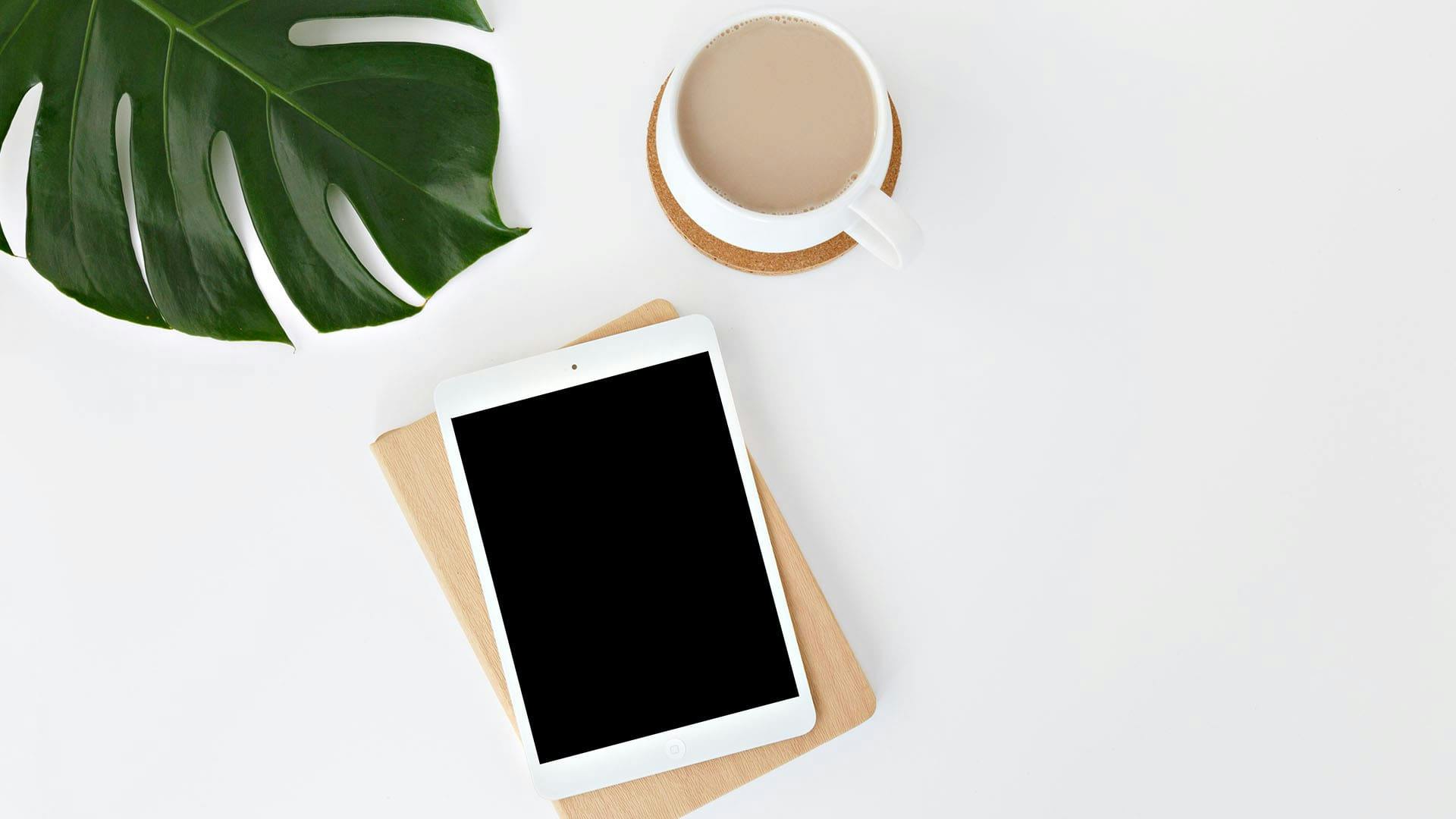 minimalist table with a white iPad on white surface, a cup of coffee and a leaf.